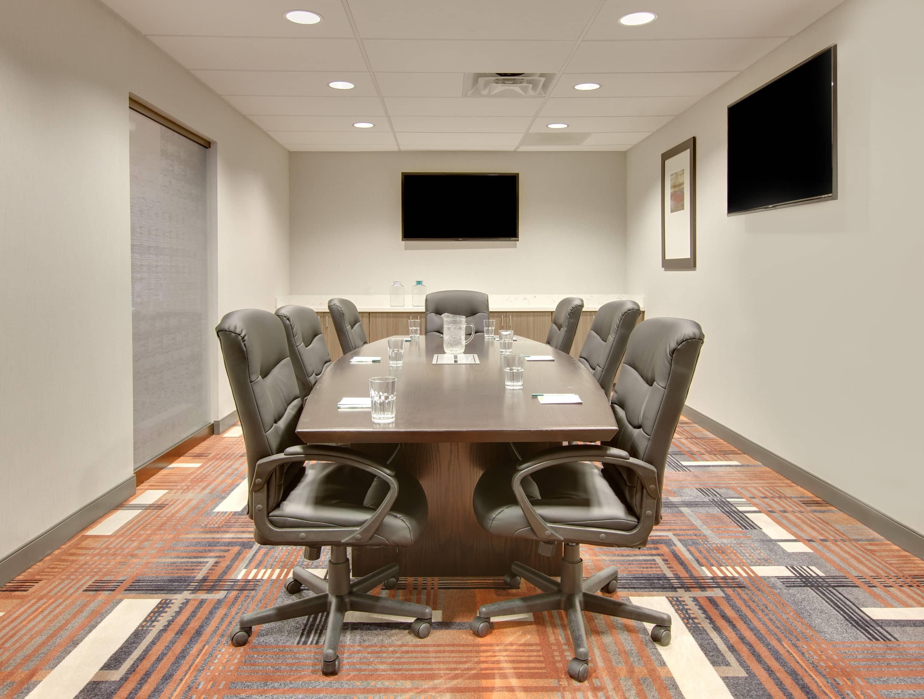Une salle de conférence avec une longue table, huit chaises, un écran de projection et un téléviseur fixé au mur.