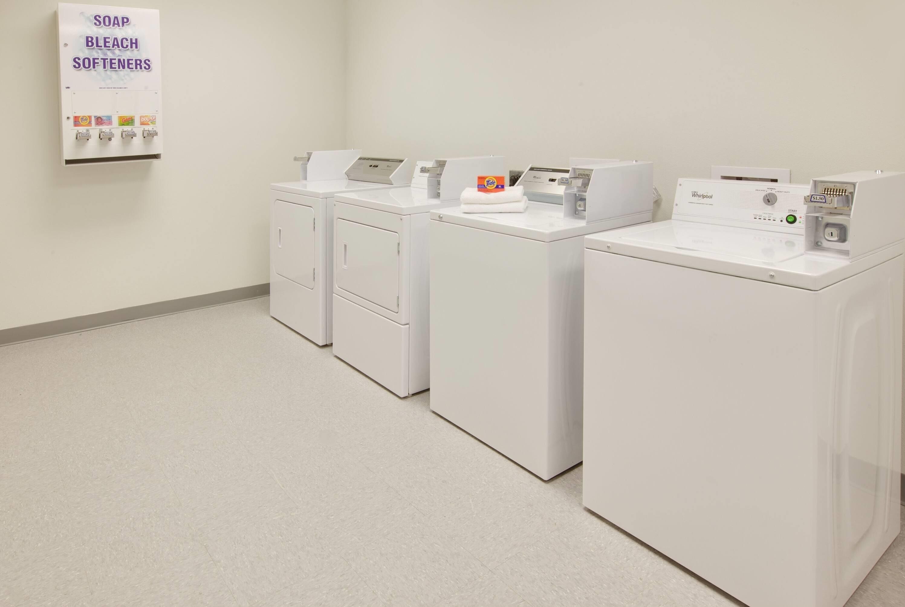 A laundry facility with four white washers and dryers, a soap, bleach, and softeners dispenser, and a Whirlpool brand dryer.