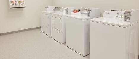 Un cuarto de lavandería con cuatro lavadoras y secadoras blancas, un dispensador de jabón, lejía y suavizantes, y una secadora Whirlpool.