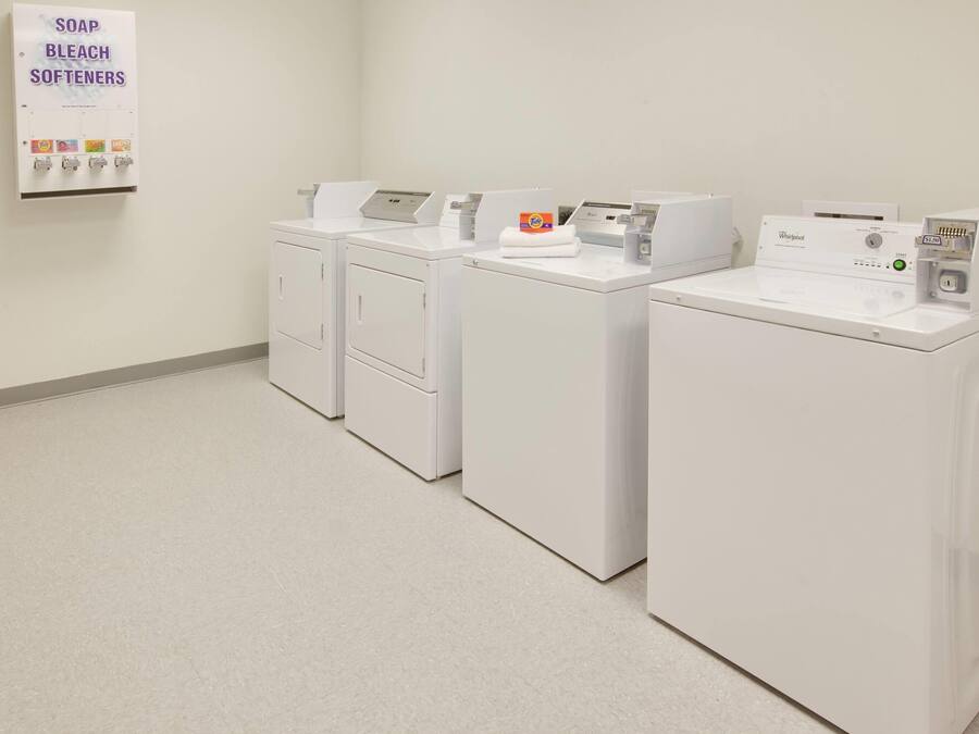 Un cuarto de lavandería con cuatro lavadoras y secadoras blancas, un dispensador de jabón, lejía y suavizantes, y una secadora Whirlpool.