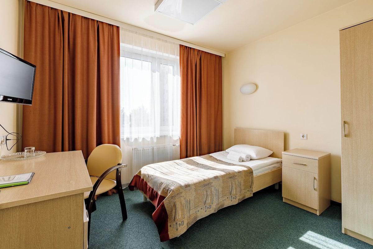 Habitación de hotel con cama, escritorio, silla, televisor, armario y ventana con cortinas.