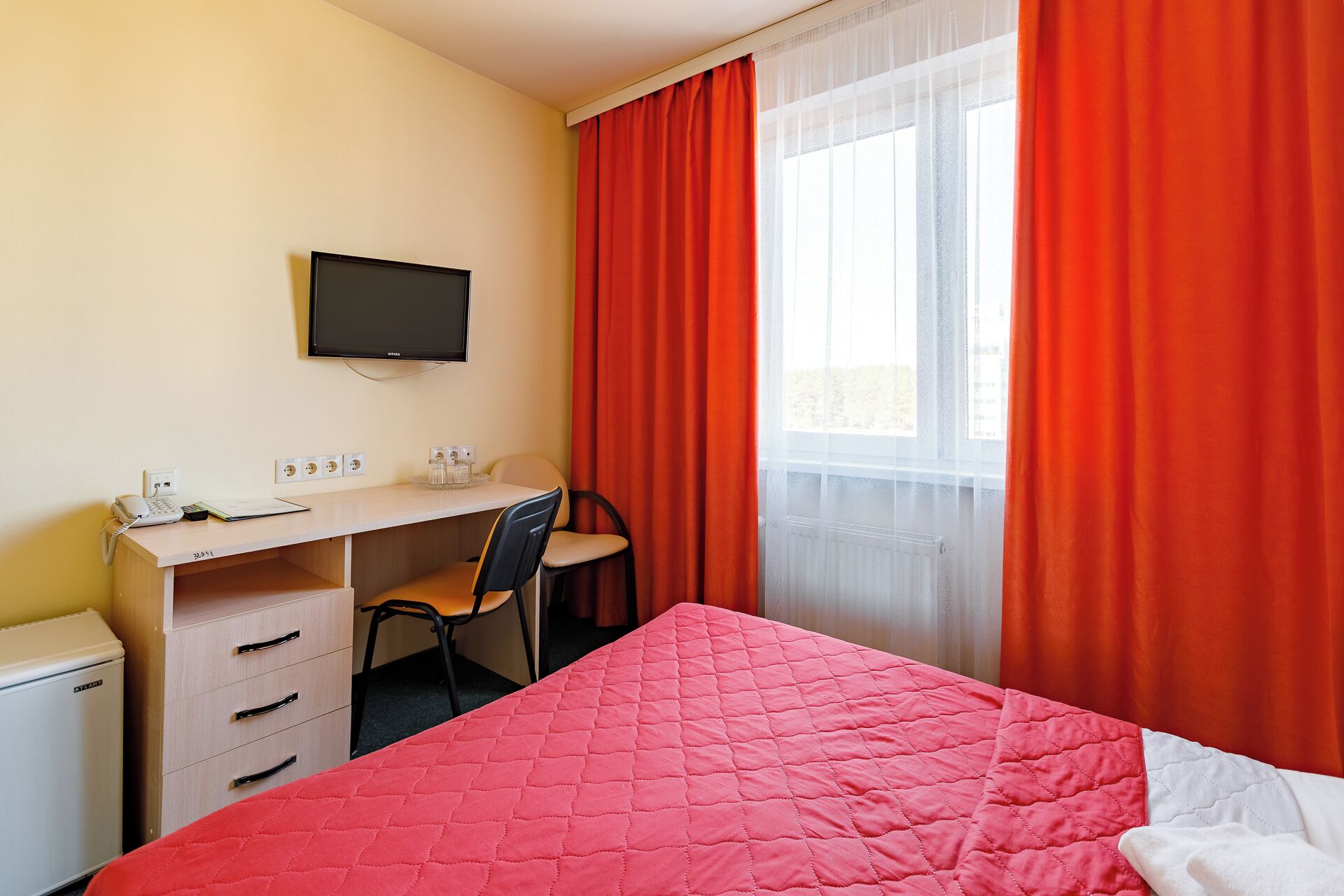 A bedroom with a bed, a desk with a chair, a television, and a window with curtains.