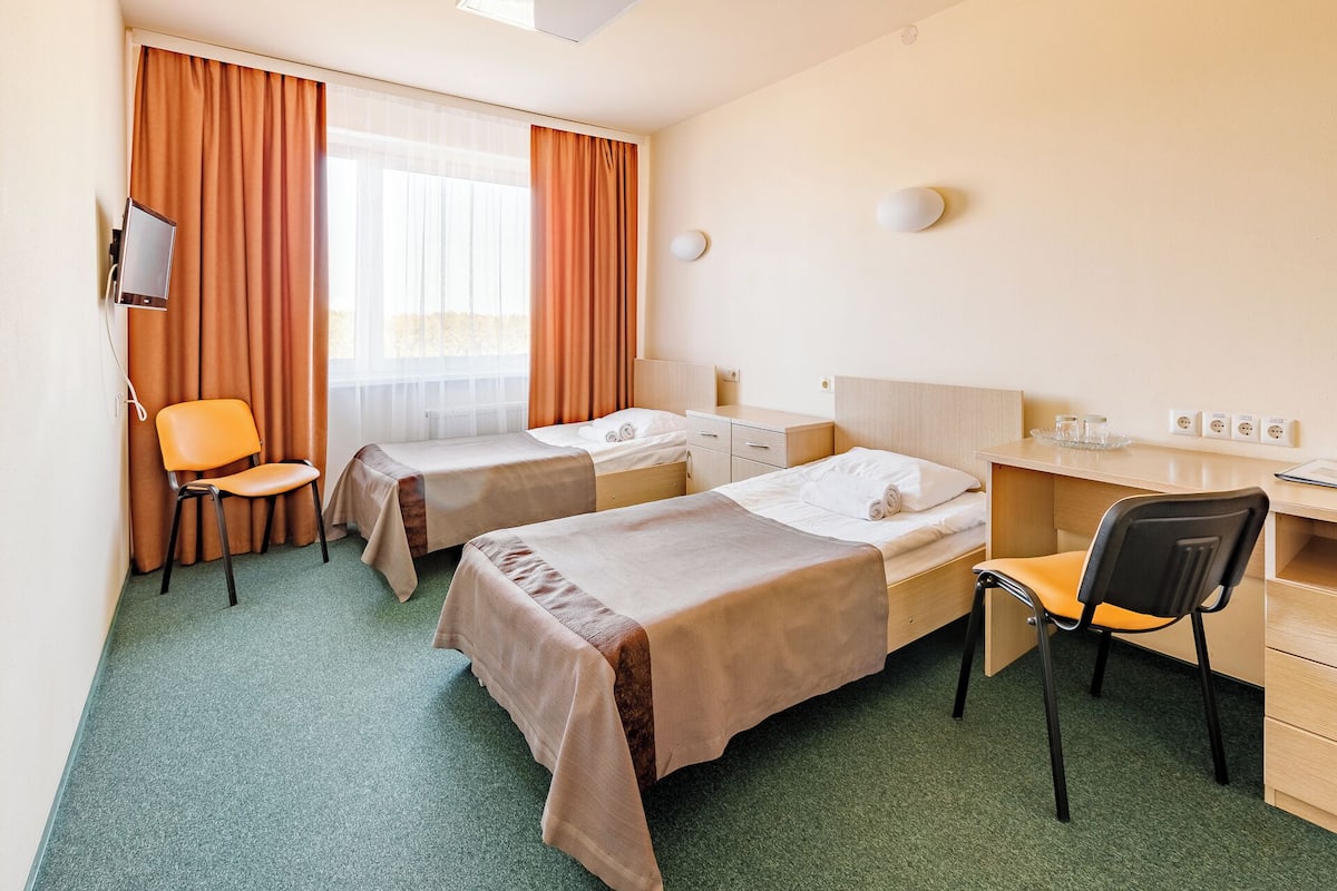 Habitación de hotel con dos camas individuales, un escritorio, una silla, un televisor y una ventana con cortinas.