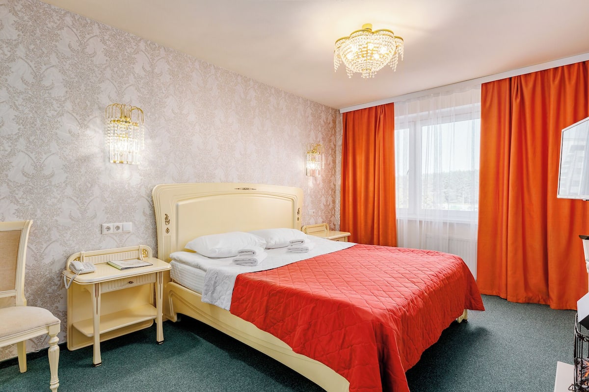 Habitación de hotel con diseño clásico, que incluye una cama con cubrecama rojo, cabecera color crema y dos lámparas de noche.