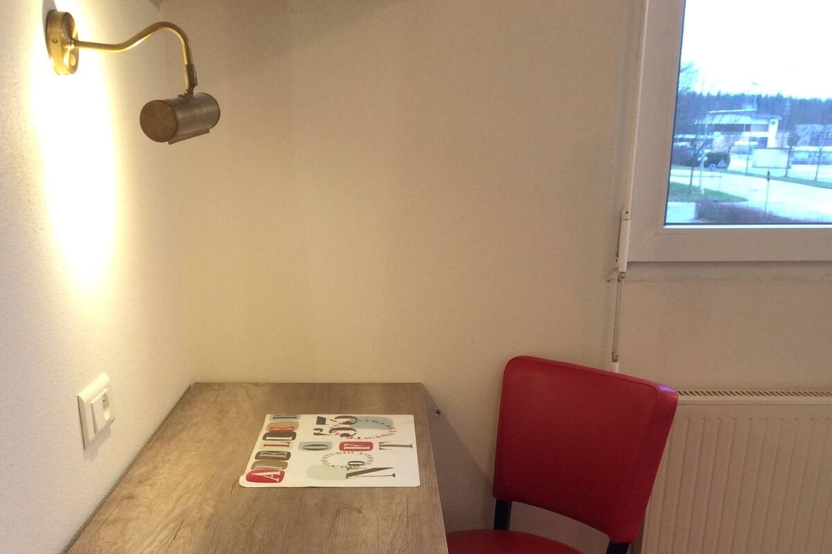 A small room with a wooden table and a red chair. There is a window with a view of a parking lot and buildings.