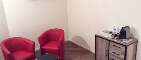 A small room with two red chairs, a small table, a coffee machine, and a cabinet with drawers.