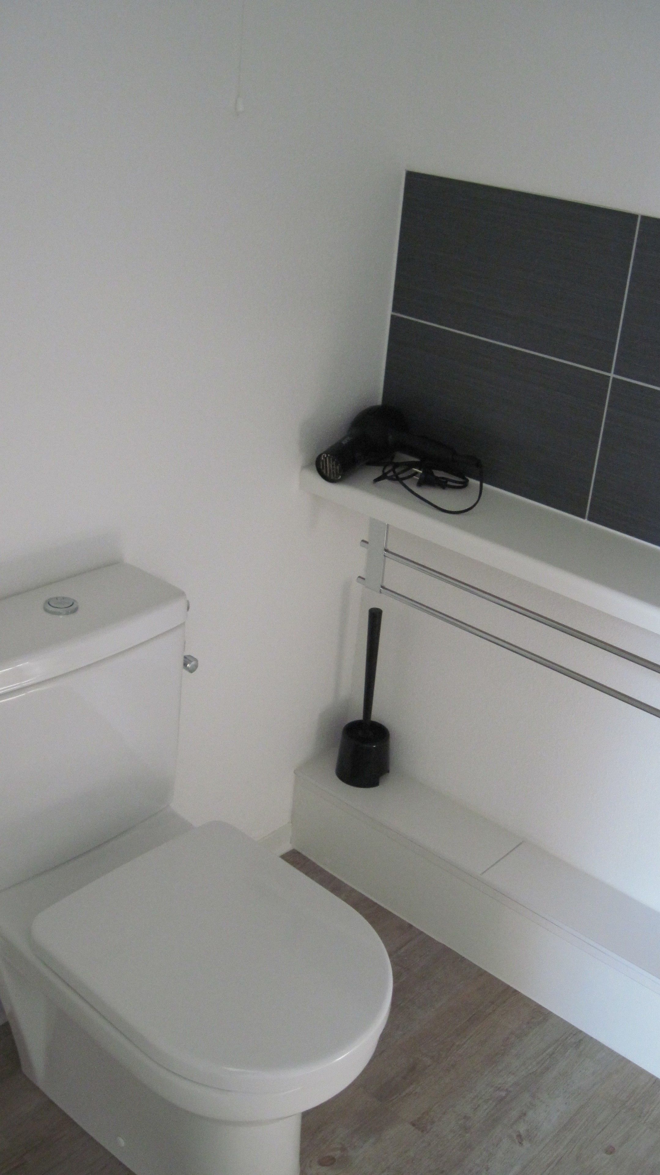 A white toilet with a closed lid, a black toilet brush, and a wall-mounted shelf with a black object on it.