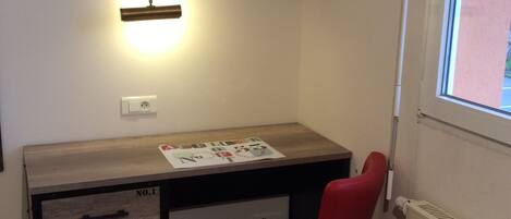 A small room with a wooden desk, a red chair, a wall-mounted TV, a shelf with a lamp, and a window.