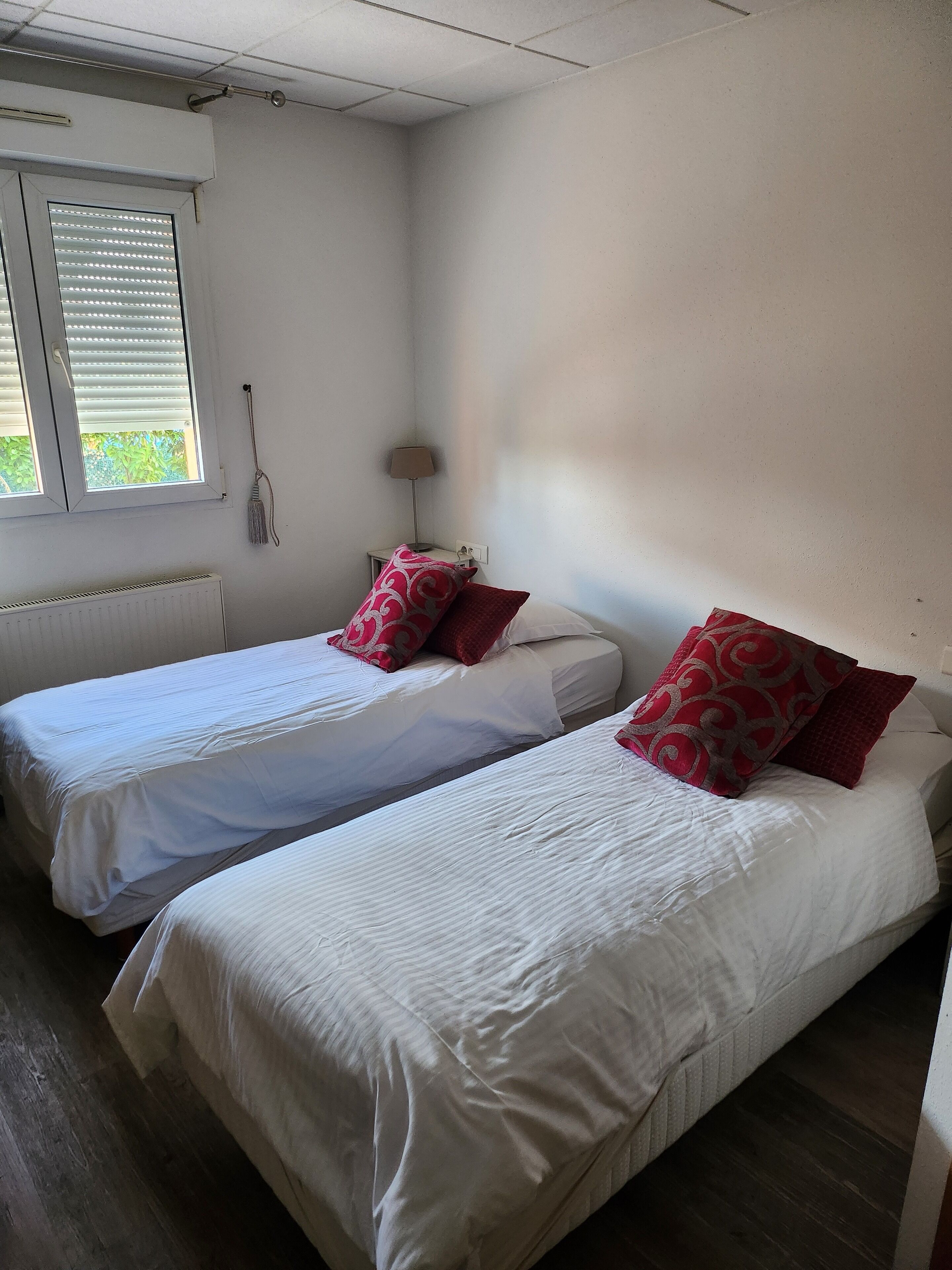 Ein Zimmer mit zwei Einzelbetten, weißer Bettwäsche und roten Kissen. Es gibt ein Fenster mit Jalousien, eine Heizung und einen kleinen Tisch mit einer Lampe.