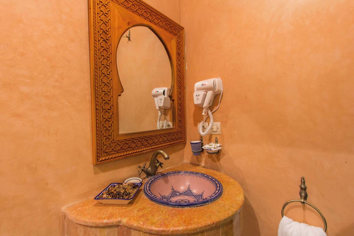 Um banheiro com uma pia redonda, um espelho com moldura decorativa e dois secadores de cabelo fixados na parede.