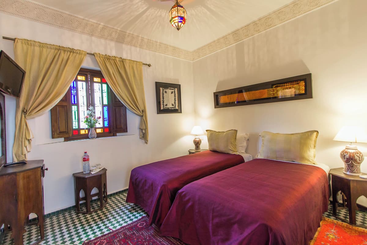 Quarto de hotel com duas camas, piso xadrez, um candelabro e uma janela com vitrais.