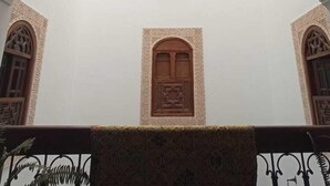 An arched entrance with a decorative lantern, a wooden door with intricate carvings, and a patterned rug in the foreground. - Dar Fes Medina Ziat (Fes)