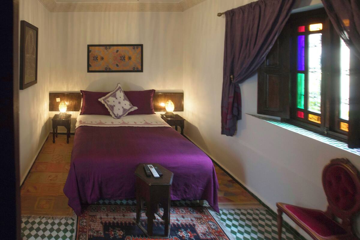 Quarto com cama, duas mesas de cabeceira, um criado-mudo, uma cadeira, janela com cortinas e um quadro na parede.