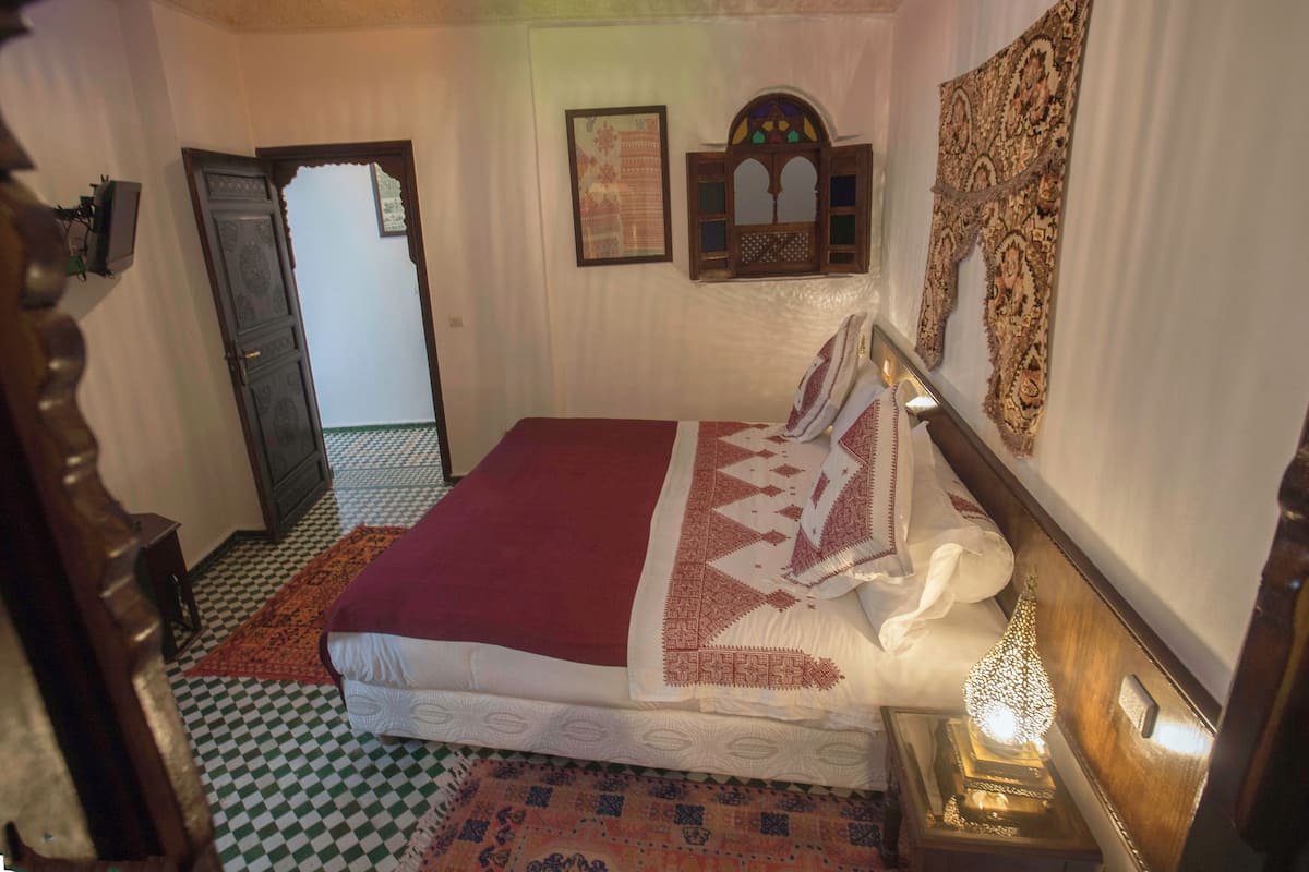 Quarto com cama, porta de madeira, tapete estampado e luminária presa na parede.