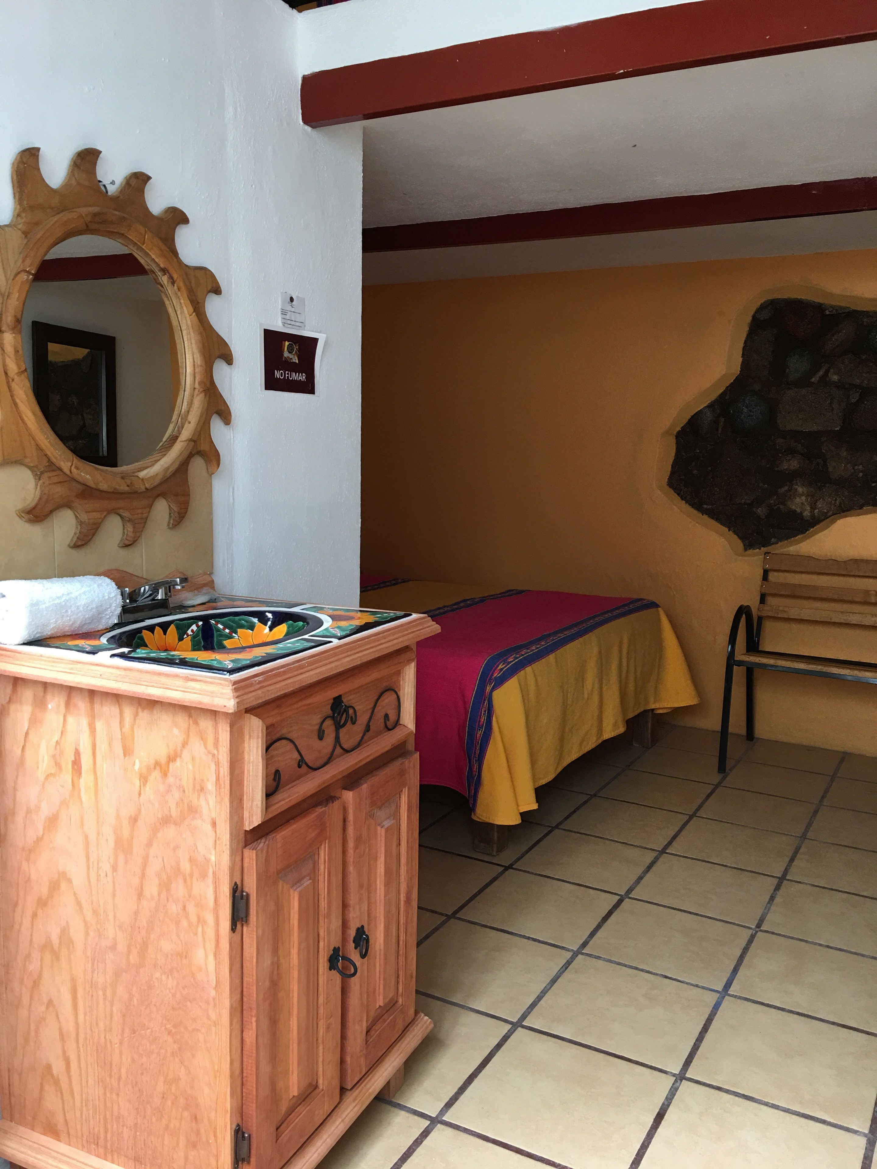 Une pièce avec un meuble en bois, un lit recouvert d’une couverture jaune et rouge, un mur en pierre et un miroir avec un cadre en bois.