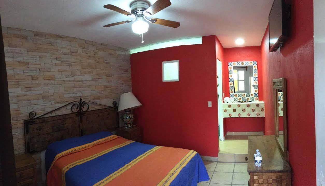 A bedroom with a bed, a ceiling fan, a nightstand, a lamp, a small window, a mirror, and a bathroom with a sink and a toilet.