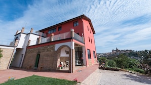 A red villa with a stone facade, a balcony, and a view of a hill with buildings. - La Casa Rossa Country House (Piazza Armerina)