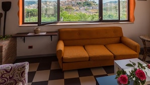 A room with a wooden ceiling, a large window with a view of a town, a sofa, a coffee table, and a patterned chair. - La Casa Rossa Country House (Piazza Armerina)