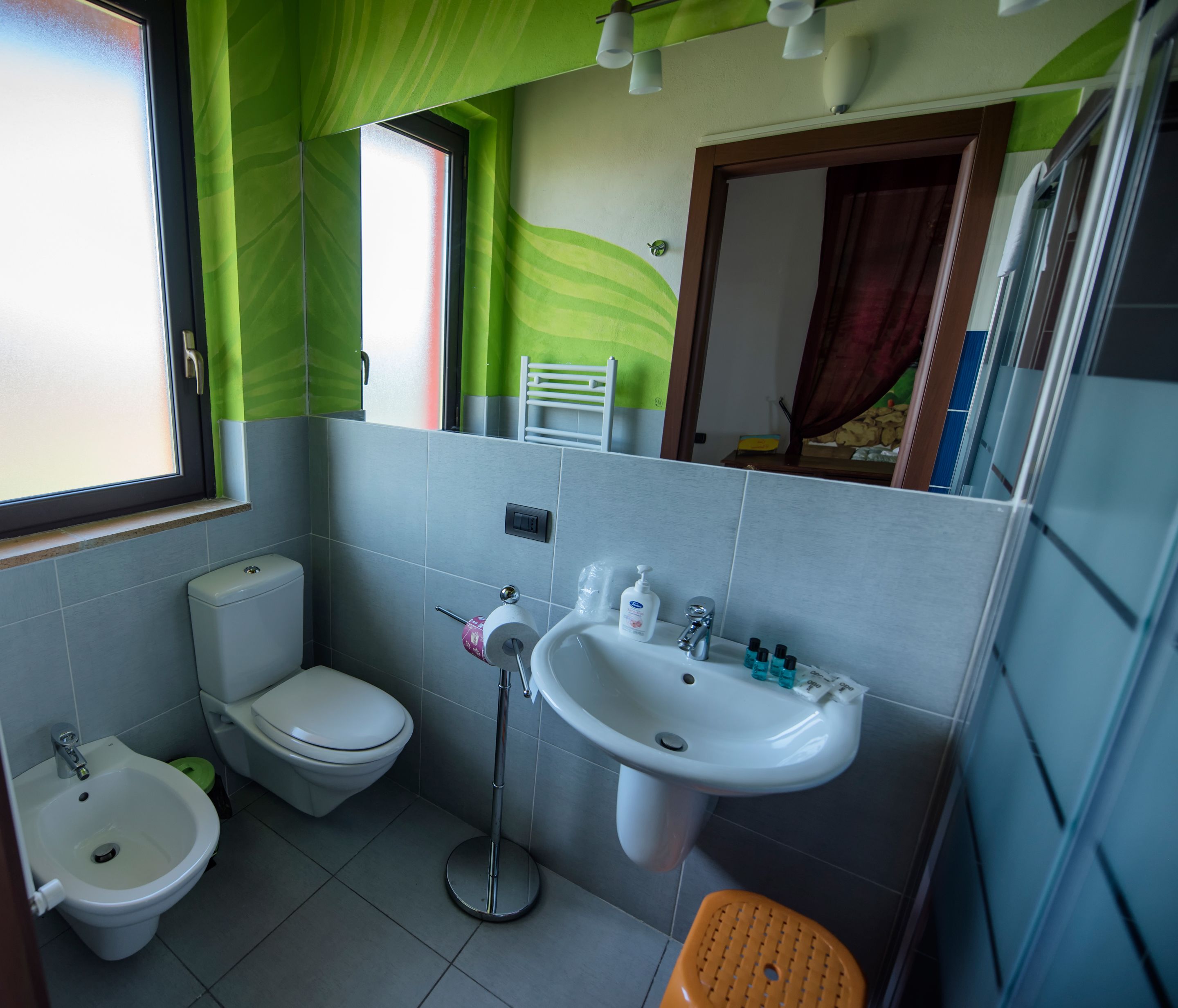 A bathroom with a bidet, toilet, sink, and a mirror reflecting a bed.