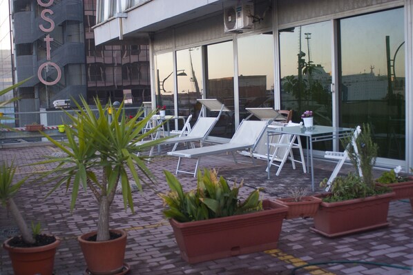An outdoor seating area with lounge chairs and tables, potted plants, and a building with a large window in the background. - Marina ResArt B&B (Naples)