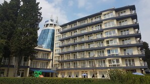 Un hotel de varios pisos con balcones, un moderno edificio de cristal a la izquierda y un jardín bien cuidado.