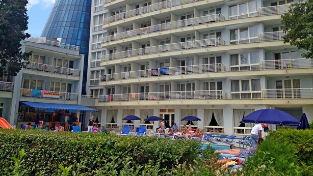 Un hotel de varios pisos con balcones, un área de piscina con tumbonas y sombrillas, y una torre azul distintiva.