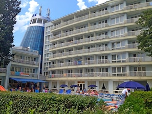A multi-story hotel with balconies, a swimming pool area with sun loungers and umbrellas, and a distinctive blue tower.