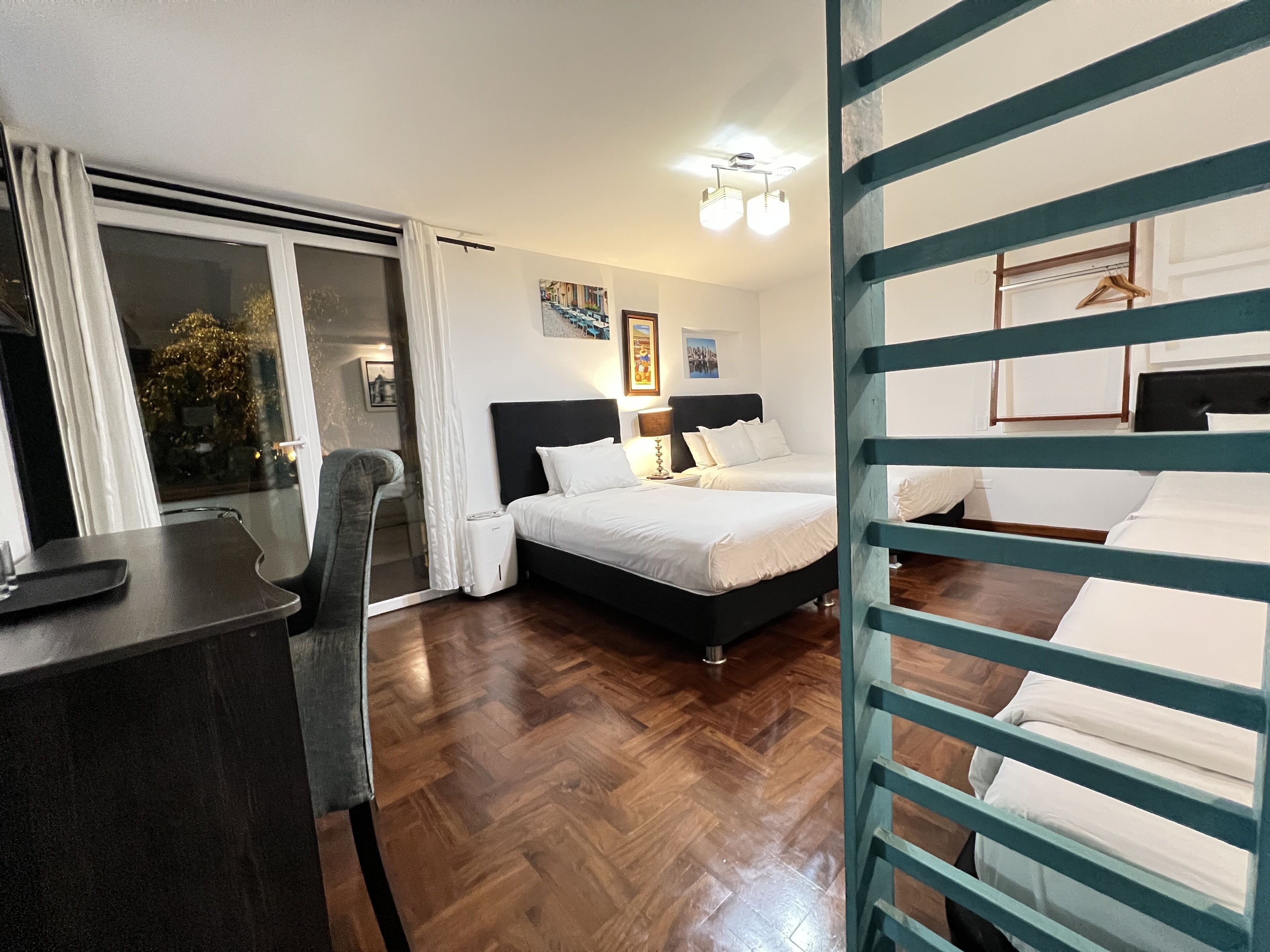Une chambre d’hôtel avec un lit, un bureau et une chaise. Il y a une fenêtre avec des rideaux, un tableau au mur et un luminaire au plafond.
