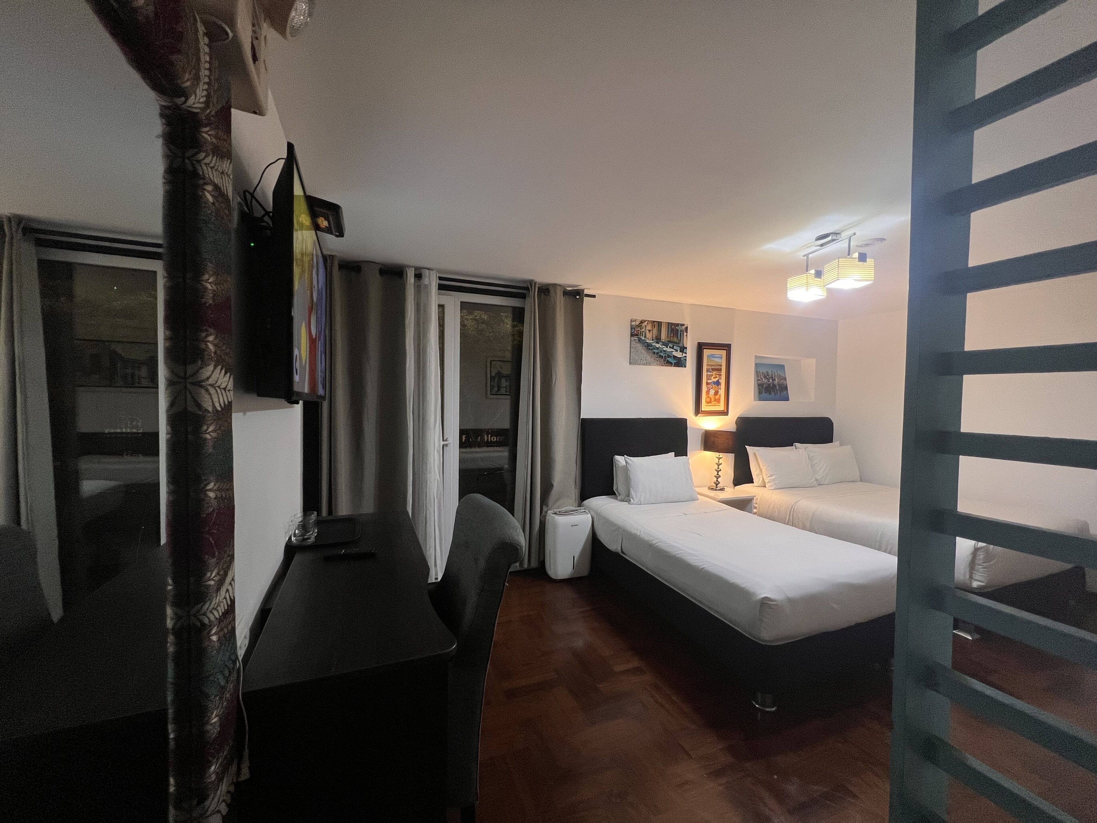 Une chambre d’hôtel avec deux lits simples, un bureau, une chaise, une petite table, une lampe et une fenêtre avec des rideaux.