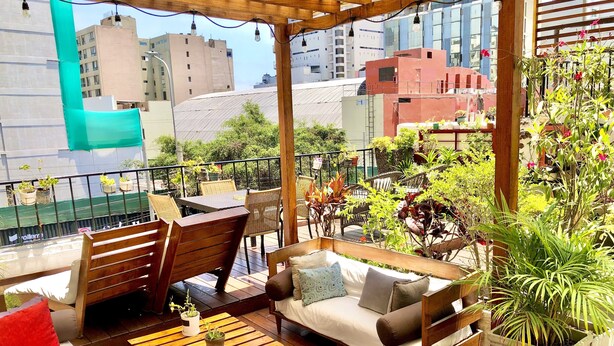 A rooftop terrace with wooden furniture, potted plants, and a view of city buildings. - La casa del Viajero (Lima)