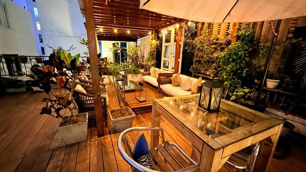 Un espacio al aire libre con piso de madera, una mesa de vidrio y un sofá con cojines.