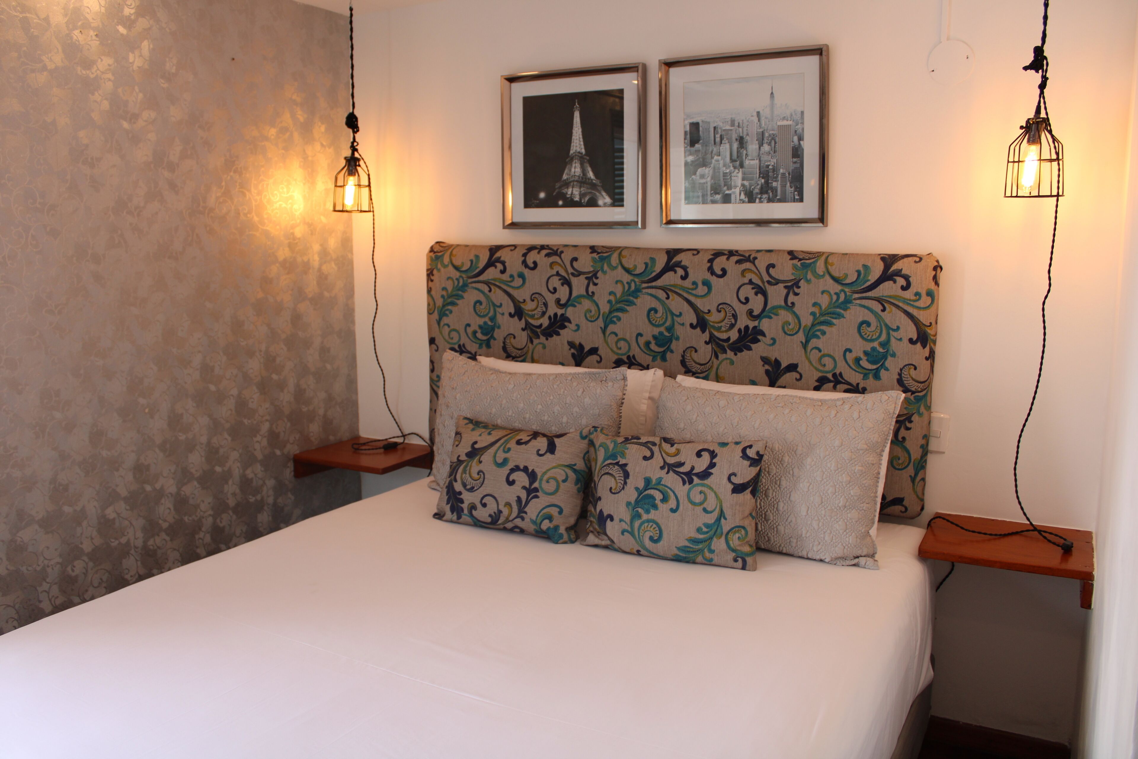 Un letto rifatto con precisione, con testiera e cuscini decorati, due quadri appesi al muro e due lampade a parete.
