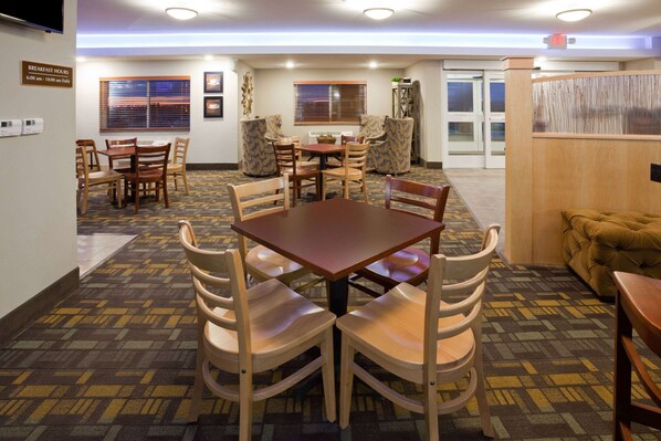 A hotel dining area with a wooden table and chairs, patterned carpet, and a sign indicating breakfast hours. - AmericInn by Wyndham Sibley (Sibley)