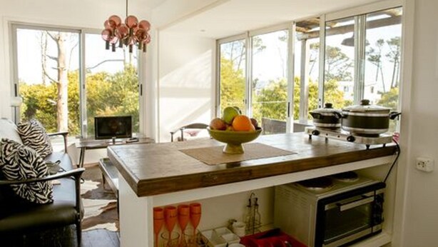 A modern kitchen with large windows, a dining area, and a living room with a leather sofa. - CDL - containers design loft (José Ignacio)