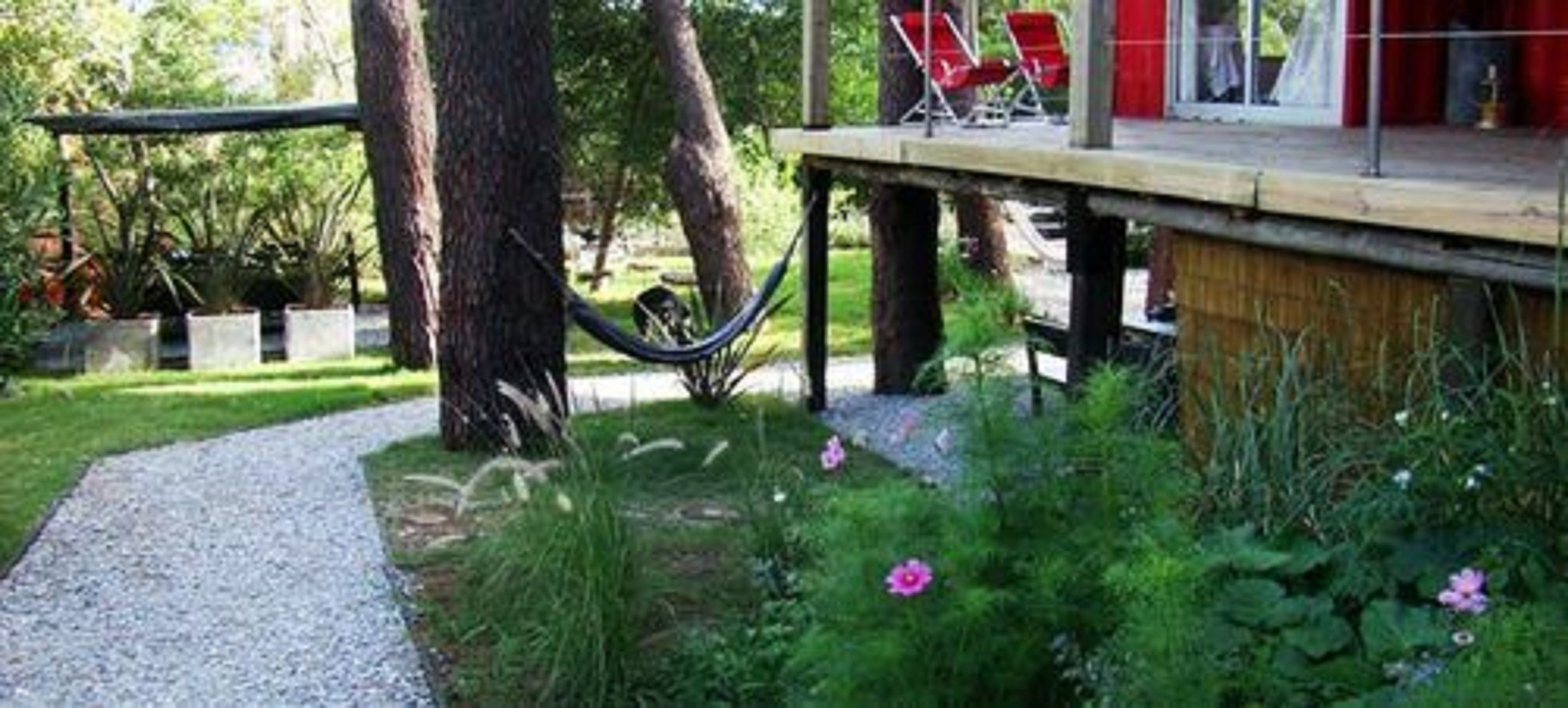 A deck with red chairs and a table, surrounded by greenery and a gravel path.