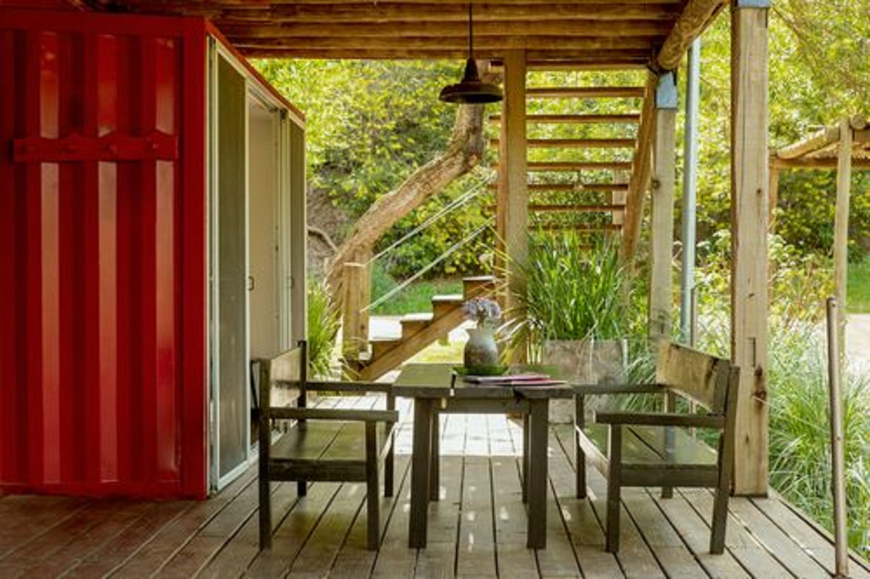 A wooden deck with a table and chairs, surrounded by greenery and a red door.