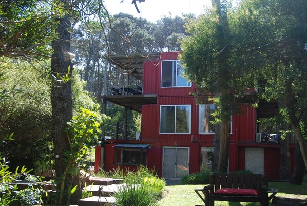 A red, two-story cabin with a deck, surrounded by trees and greenery.