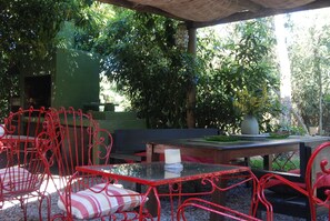 Un área de estar al aire libre con sillas metálicas rojas y una mesa de vidrio, rodeada de follaje y un banco de madera.