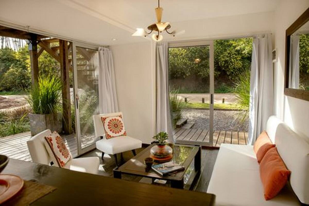 A modern living room with a sofa, a coffee table, and a chair. There is a large window with curtains, a ceiling fan, and a view of a garden.