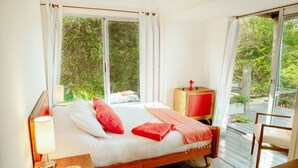 A bedroom with a large bed, a nightstand, a bedside table, a red lamp, and a view of greenery outside. - CDL - containers design loft (José Ignacio)