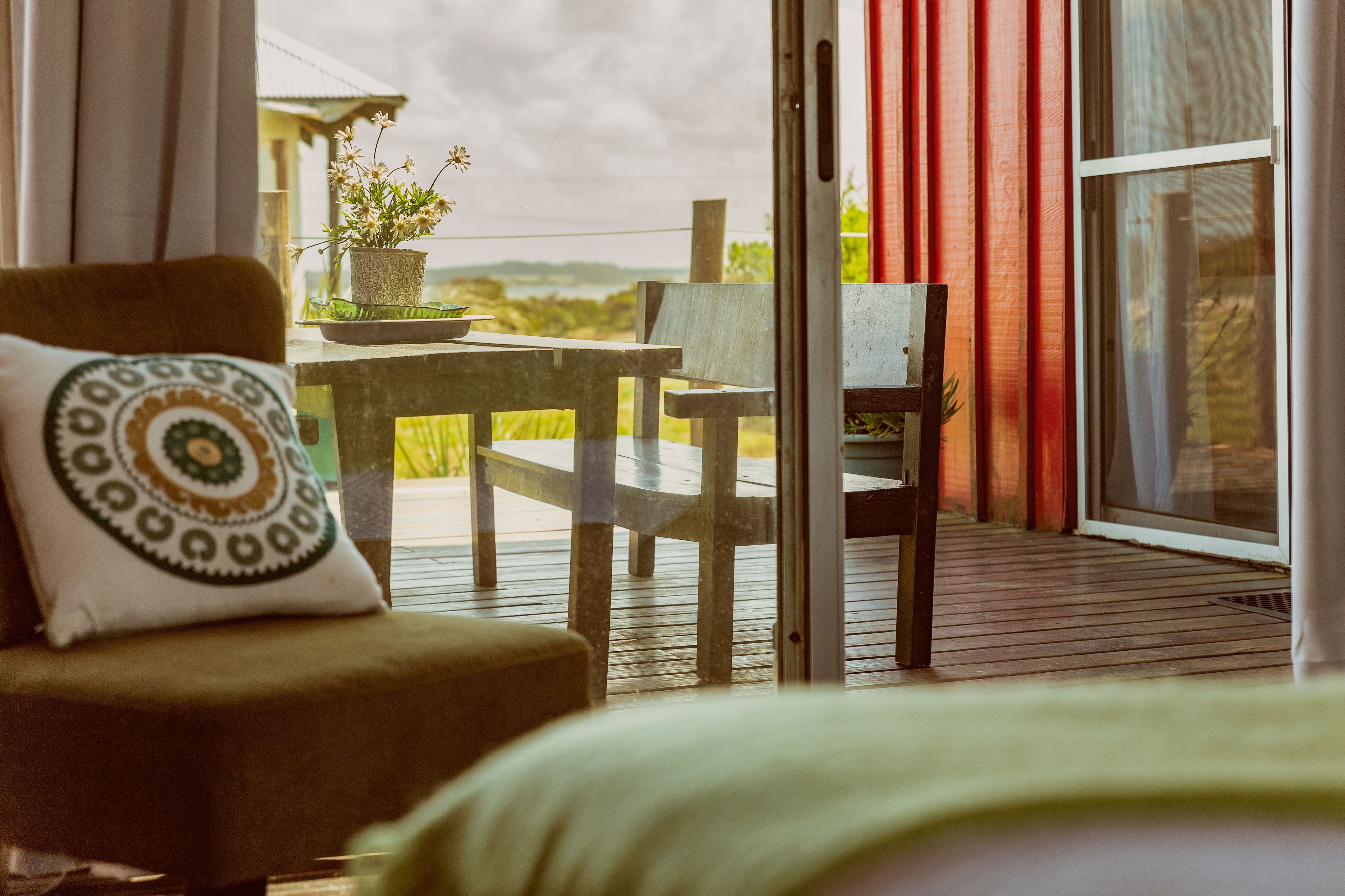 A room with a bed, a chair with a patterned pillow, a table with a potted plant, and a view of the outdoors through a sliding door.
