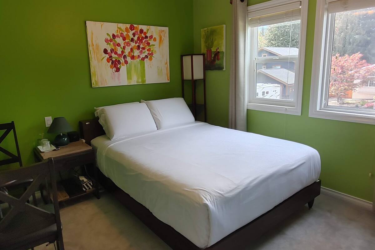 Un dormitorio con cama, una silla, una mesita de noche, una lámpara y un cuadro en la pared.