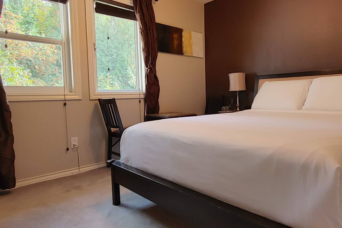 Una cama bien hecha en una habitación de hotel, con un ventanal grande, una silla y una mesita de noche con lámpara.