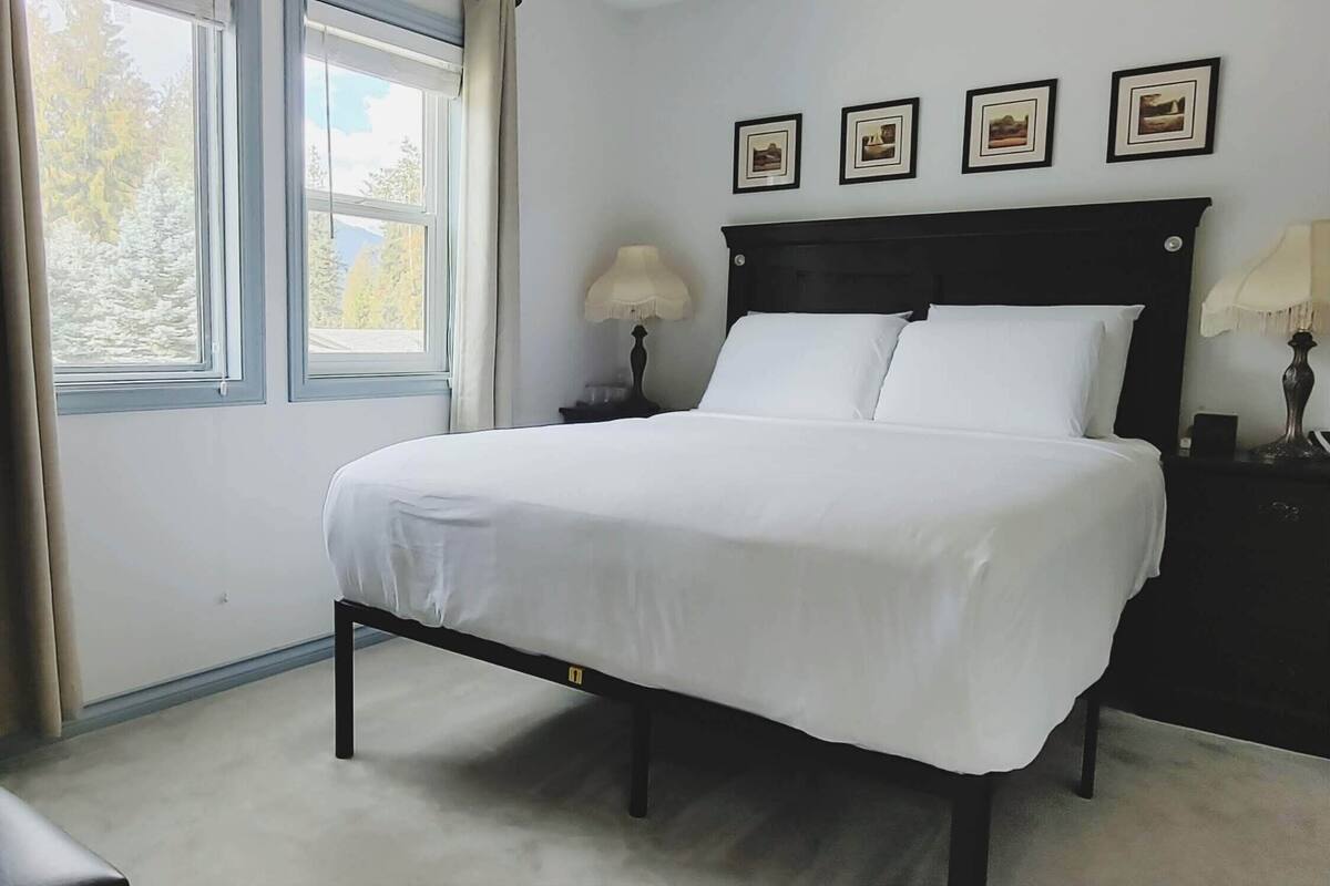 Una cama bien hecha en una habitación con ventana, una mesita de noche con lámparas y cuadros en la pared.