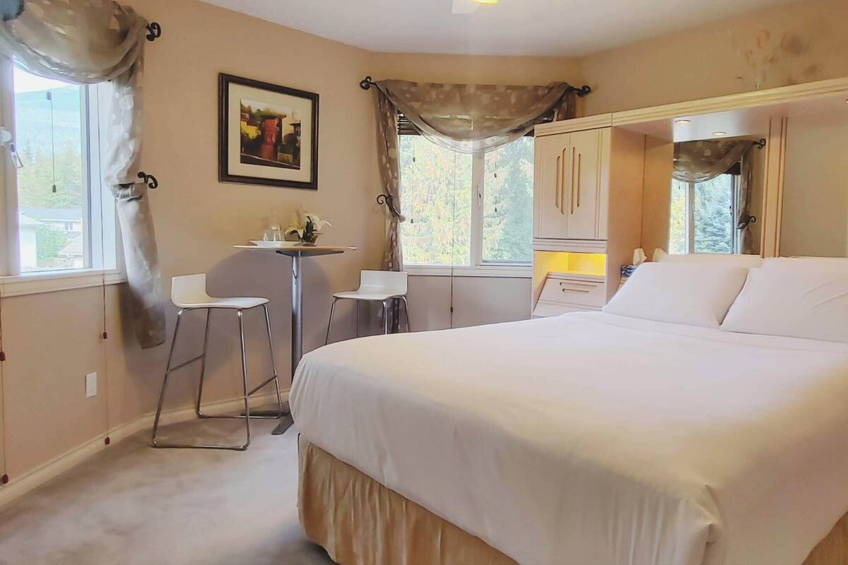 Un dormitorio con una cama grande, un escritorio y una silla. Hay una ventana con cortinas, un cuadro en la pared y un espejo.