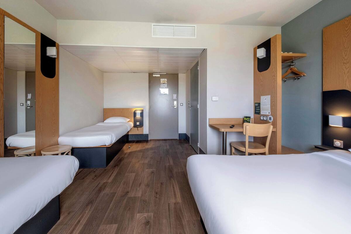 Habitación de hotel con dos camas individuales, un escritorio con silla, una mesita, un espejo y un estante en la pared.