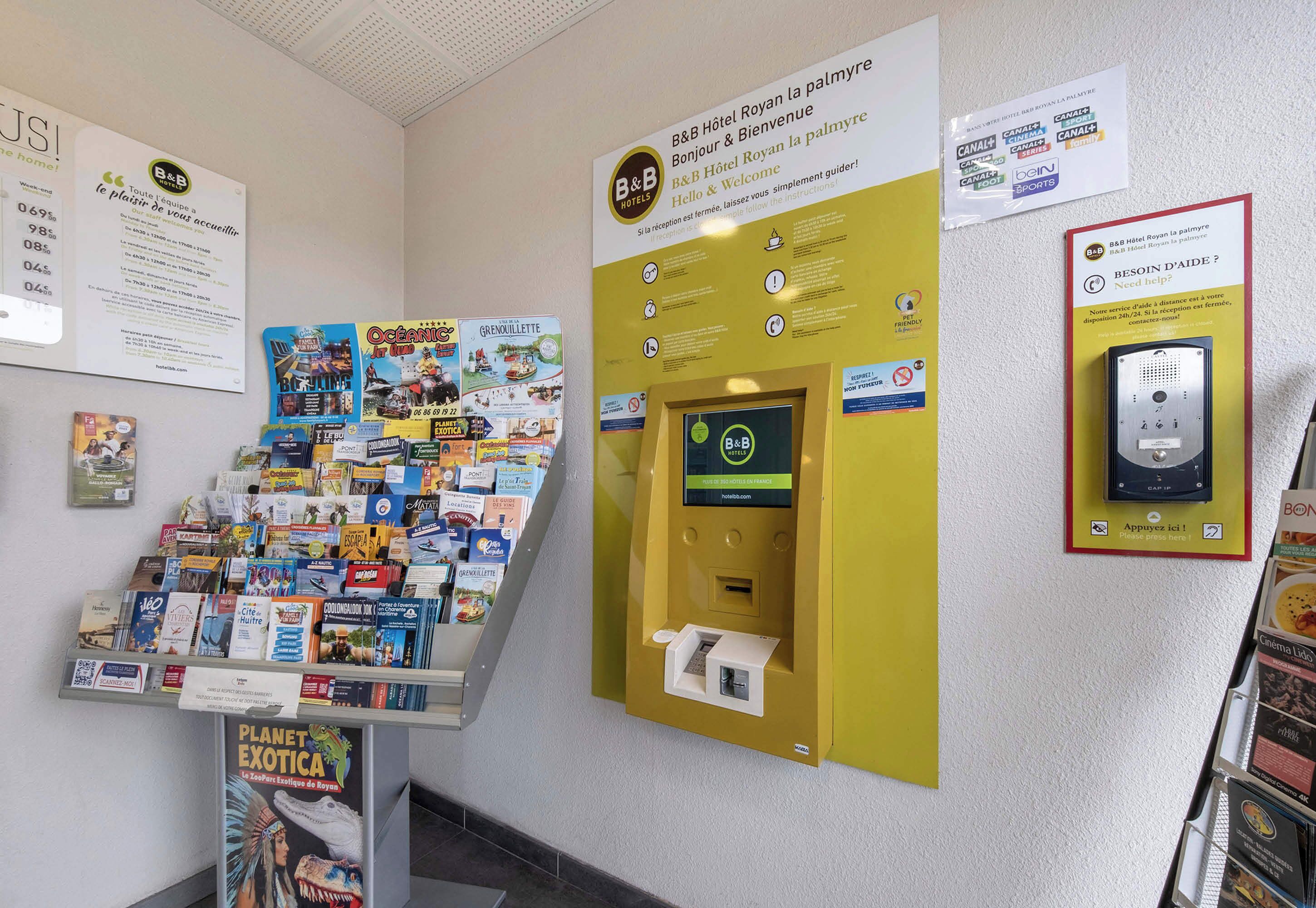 Un espace d’accueil d’hôtel comprenant un kiosque en libre-service, des affiches d’information et un présentoir de brochures de voyage.