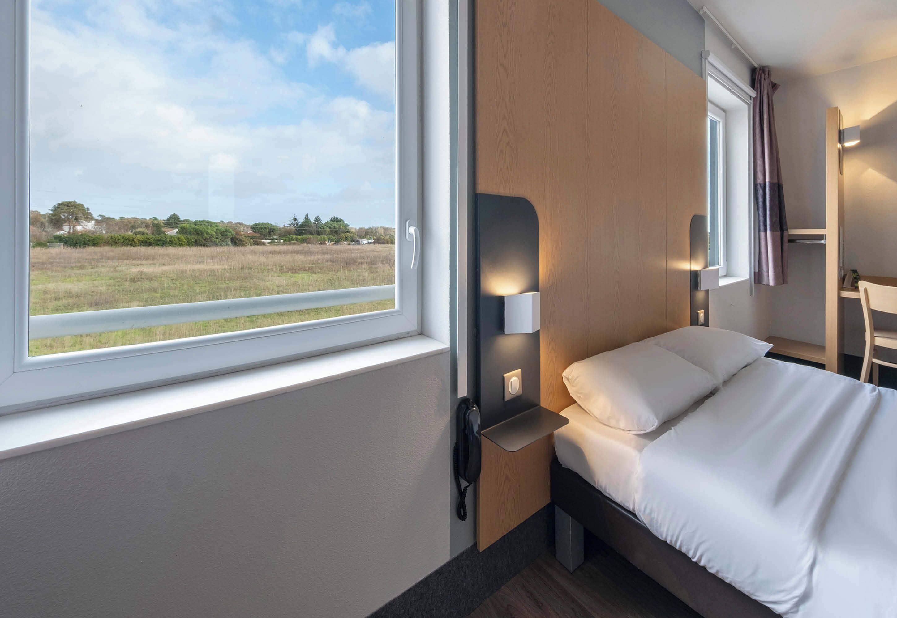 Ett hotellrum med en säng, ett sängbord, en lampa, ett fönster med utsikt över ett fält och en dörr.