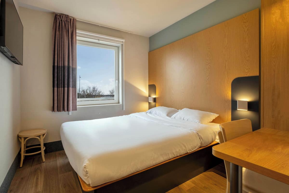 Habitación de hotel con cama, una mesita, una silla, televisor y ventana con cortinas.