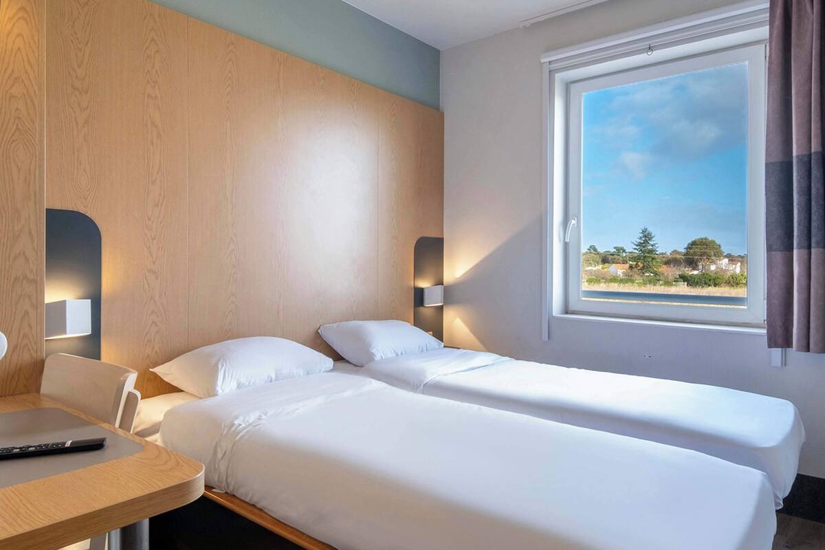Habitación de hotel con dos camas individuales, un escritorio y una ventana con vistas al paisaje.